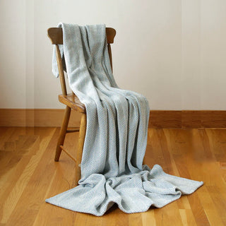A light blue and white herringbone patterned throw blanket is draped over a rustic wooden chair in a room with light beige walls and a polished wood floor.