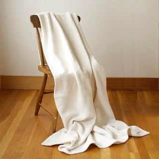 A cream-colored textured woven cotton throw blanket draped over a wooden chair in a minimalist room with light wood floors and a beige wall.