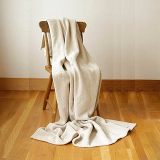 A cream and beige herringbone textured cotton throw draped on a chair with a cream wall and warm wood floor.