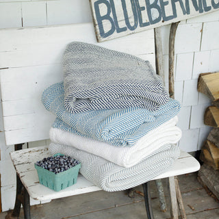 A stack of folded woven textiles in neutral and blue tones sits on a white wooden bench, with a small basket of blueberries on the front and a rustic sign in the background.