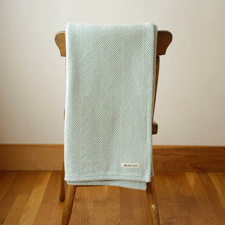 A folded pale mint and natural herringbone patterned throw blanket draped over a wooden chair in a room with a beige wall and wooden floor.