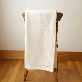 A natural colored cotton throw in a herringbone texture, folded neatly and draped over a chair in a room with cream wall and warm oak floor.  