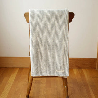 A light grey and white herringbone patterned throw blanket draped over a wooden chair in a room with light wood floors and a cream-colored wall.