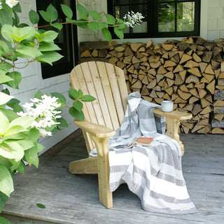 A light natural wood Adirondack chair sits on a weathered gray porch deck, draped with a gray and white striped woven cotton throw blanket. A small wooden side table on the chair holds a white mug, and a stack of firewood is visible in the background against a white house wall with green leaves in the foreground.