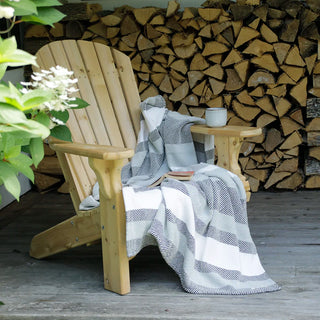 A light natural wood Adirondack chair on a wooden deck with a gray and white striped cotton throw blanket draped over it, set against a stacked wood firewood backdrop.