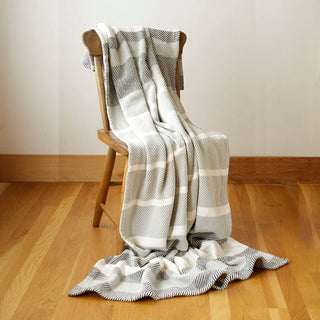 A gray and white striped cotton throw blanket with a textured woven pattern and hemmed edges, draped over a wooden chair on a light wood floor against a neutral wall.