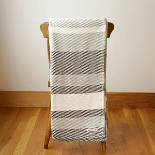 A striped woven cotton blanket in shades of light gray, white, and dark grey is draped over the back of a wooden chair in a room with light wood floors and a beige wall.