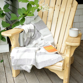 A light natural wood Adirondack chair on a weathered wooden deck, styled with a dark grey and white striped cotton throw blanket, a white mug on the armrest, and a book.