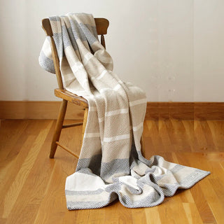 A cream, beige,  and gray striped throw blanket draped over a wooden chair on a light wood floor against a neutral wall.